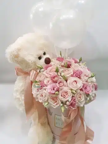 Caja decorativa con flores frescas de colores vibrantes y un peluche de oso, ideal para regalar en ocasiones especiales.