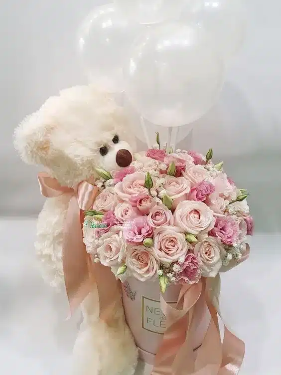 Caja decorativa con flores frescas de colores vibrantes y un peluche de oso, ideal para regalar en ocasiones especiales.